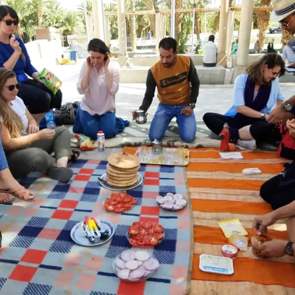 Picnic lunch in Midelt on the way from the City of Fes to the Sahara desert. The group are sharing a lunch in an oasis with colourful blankets on the ground, sampling a Moroccan style picnic and experiencing Moroccan hospitality on the road. Morocco Explored