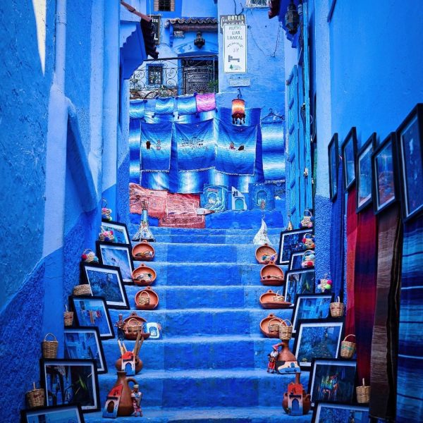 One of the very blue narrow staircases in the town of Chefchaouen, the steps lead up to an archway at the top and have a selection of blankets and Moroccan handicrafts on the walls and each step, Morocco Uncovered
