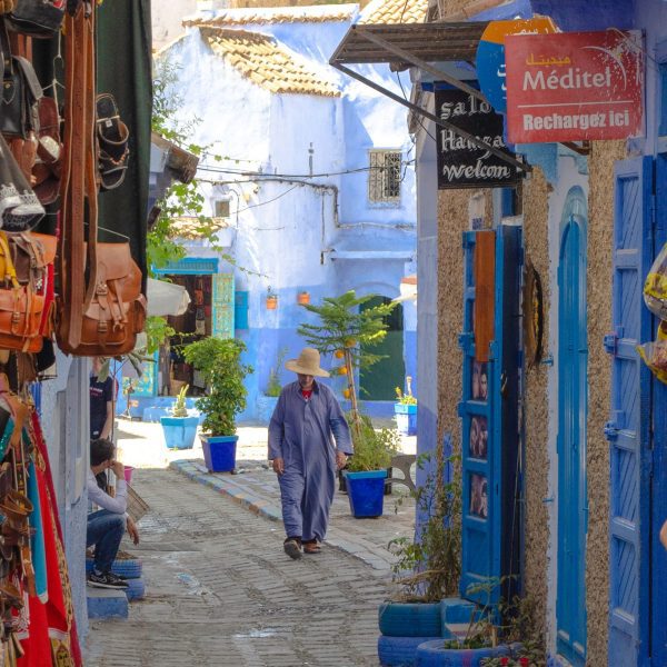 Local man in traditional costume walking through a narro street in the medina of the Blue city, Chefchaouen. There are shops on either side of the street selling local handicrafts. Classic Morocco. Morocco Explored